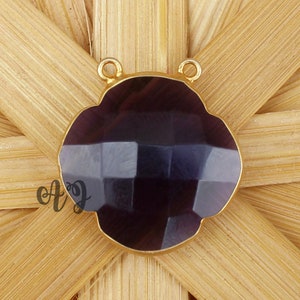 Può includere: Un ciondolo dorato con una pietra preziosa di colore viola scuro a forma di fiore. Il ciondolo ha due anelli nella parte superiore per attaccare una catena.