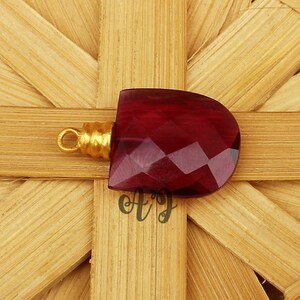 May include: A deep red faceted gemstone pendant with a gold-toned bail. The pendant is on a woven background.