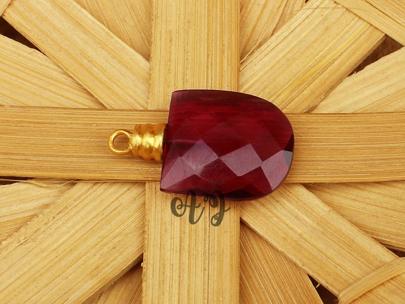 May include: A deep red faceted gemstone pendant with a gold-toned bail. The pendant is on a woven background.
