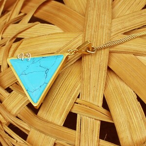 May include: A gold-toned necklace with a triangular pendant featuring a vibrant turquoise stone. The pendant has the initials "AJ" in a delicate script. The necklace is displayed on a woven, light brown surface.