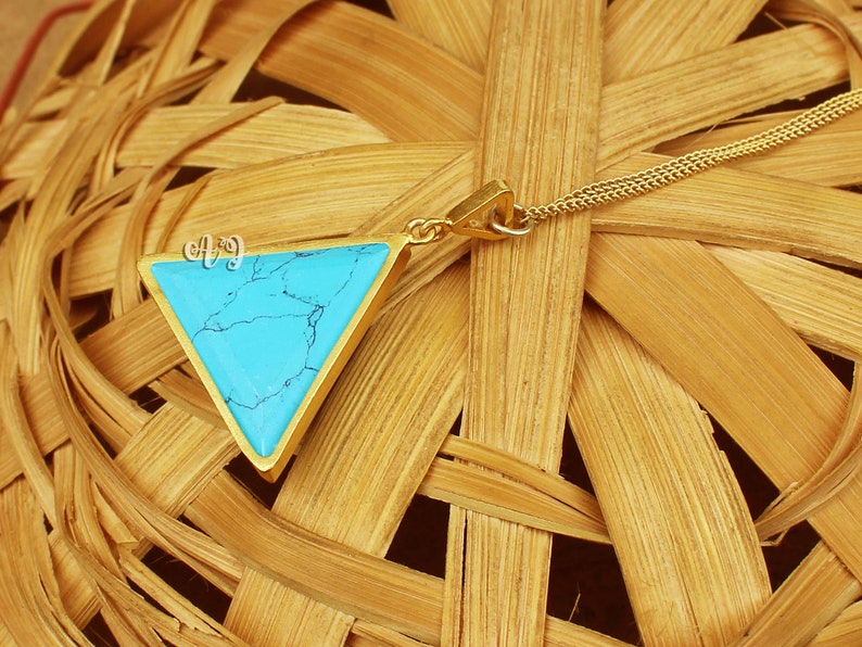 May include: A gold-toned necklace with a triangular pendant featuring a vibrant turquoise stone. The pendant has the initials "AJ" in a delicate script. The necklace is displayed on a woven, light brown surface.