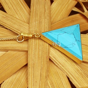 May include: A gold-colored necklace featuring a triangular turquoise pendant. The pendant has a gold border and a chain. The turquoise stone has a marbled appearance with dark veins. The necklace is displayed on a woven background.
