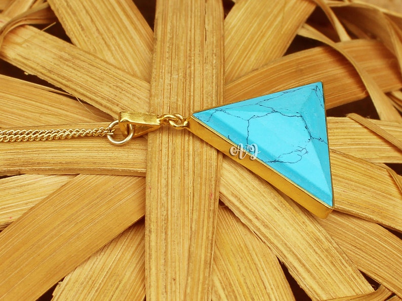 May include: A gold-colored necklace featuring a triangular turquoise pendant. The pendant has a gold border and a chain. The turquoise stone has a marbled appearance with dark veins. The necklace is displayed on a woven background.
