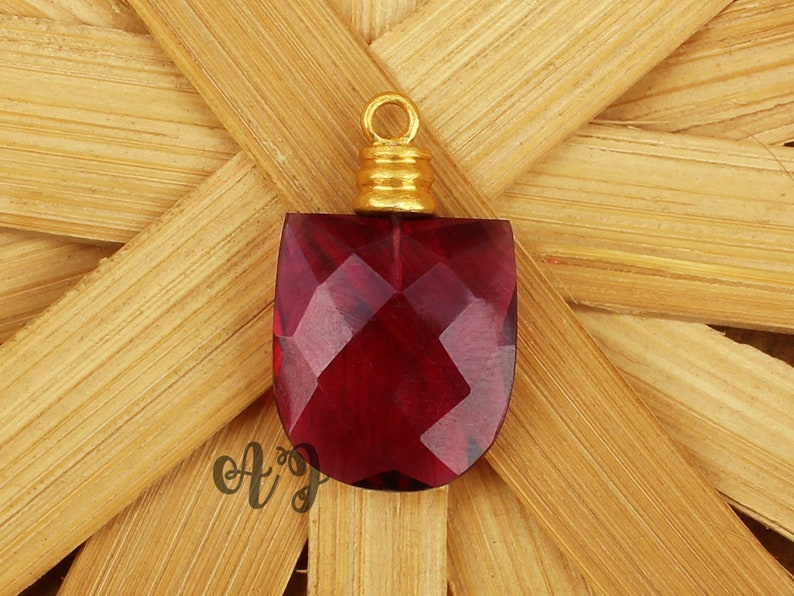 May include: A deep red faceted glass pendant with a gold-toned metal bail. The pendant is shaped like a shield and has a faceted design.