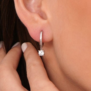 May include: Silver hoop earrings with a small, clear gemstone dangling from the bottom. The hoop is lined with small, clear gemstones.