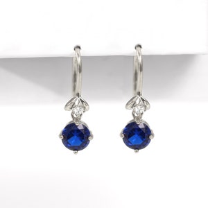 Peut inclure: Une paire de boucles d'oreilles créoles argentées. Chaque boucle d'oreille présente une petite pierre précieuse transparente au-dessus d'une pierre précieuse ronde bleu foncé. Les boucles d'oreilles sont présentées sur fond blanc.