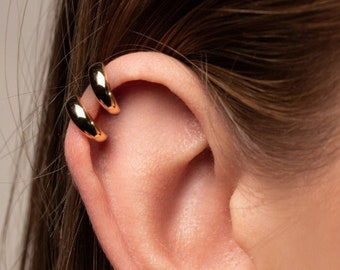 May include: Close-up of an ear adorned with two gold-colored ear cuffs. The ear cuffs are smooth, rounded, and hug the ear's cartilage. The image showcases a minimalist jewelry style.