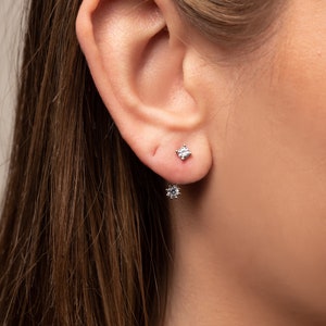 Peut inclure: Gros plan sur une oreille avec deux petites boucles d'oreilles rondes et transparentes. Les boucles d'oreilles sont en argent et ont une seule pierre transparente au centre.