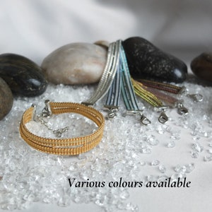 Peut inclure: Un bracelet en perles dorées avec un fermoir argenté. Le bracelet est composé de plusieurs rangs de perles et est présenté sur une surface blanche avec de petites pierres claires.  Différentes couleurs disponibles.