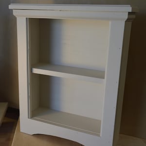 May include: A white wooden bookshelf with one shelf. The bookshelf has a simple design with rounded edges and a slightly curved base.