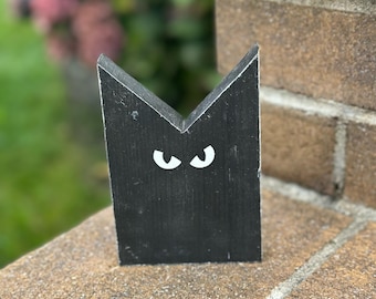 May include: Black wooden cat cutout with white eyes. The cat is standing on a brown brick surface.
