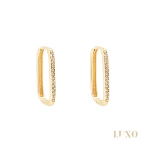 Peut inclure: Une paire de boucles d'oreilles créoles rectangulaires dorées. Chaque boucle d'oreille est ornée de petits cristaux transparents sur un côté. Les boucles d'oreilles sont présentées sur fond blanc. Le mot "LUXO" est imprimé en bas à droite.