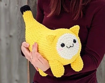 May include: A crocheted banana-shaped plush toy with a white face and black eyes. The toy is being held by a person wearing a maroon shirt.