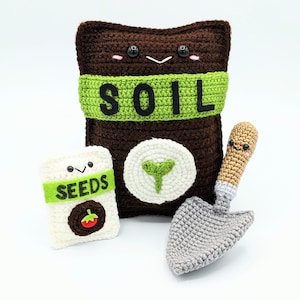 May include: A brown crocheted bag with the word "SOIL" on it, a white crocheted bag with the word "SEEDS" on it, and a grey crocheted trowel with a brown handle.