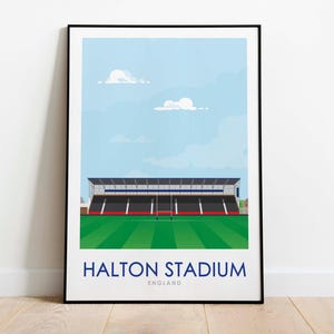 Può includere: Una stampa incorniciata nera di un'illustrazione di cartoni animati dello stadio di Halton in Inghilterra. Lo stadio è verde con un tetto bianco e blu. Il cielo è blu con nuvole bianche.