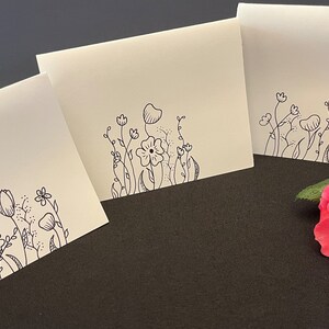 Puede incluir: Tres tarjetas blancas en blanco con dibujos de flores y hojas en tinta negra en el borde inferior. Las tarjetas est&aacute;n sobre una superficie gris oscuro con una rosa rosa en el lado derecho de la imagen.