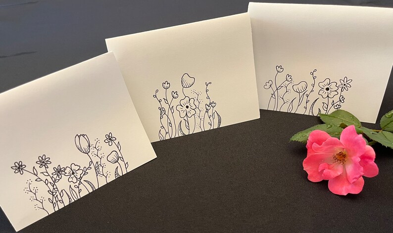 Puede incluir: Tres tarjetas blancas en blanco con dibujos de flores y hojas en tinta negra en el borde inferior. Las tarjetas est&aacute;n sobre una superficie gris oscuro con una rosa rosa en el lado derecho de la imagen.
