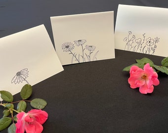 Puede incluir: Tres tarjetas de felicitación color crema con dibujos lineales florales negros. Una tarjeta presenta una sola margarita, otra tiene tres margaritas y la tercera tiene una variedad de flores. Una rosa rosa y hojas están en primer plano.