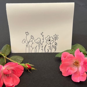 Puede incluir: Una tarjeta blanca en blanco con un dibujo de tinta negra de una fila de flores y tallos simples. La tarjeta est&aacute; sobre una superficie negra con una rosa rosa.