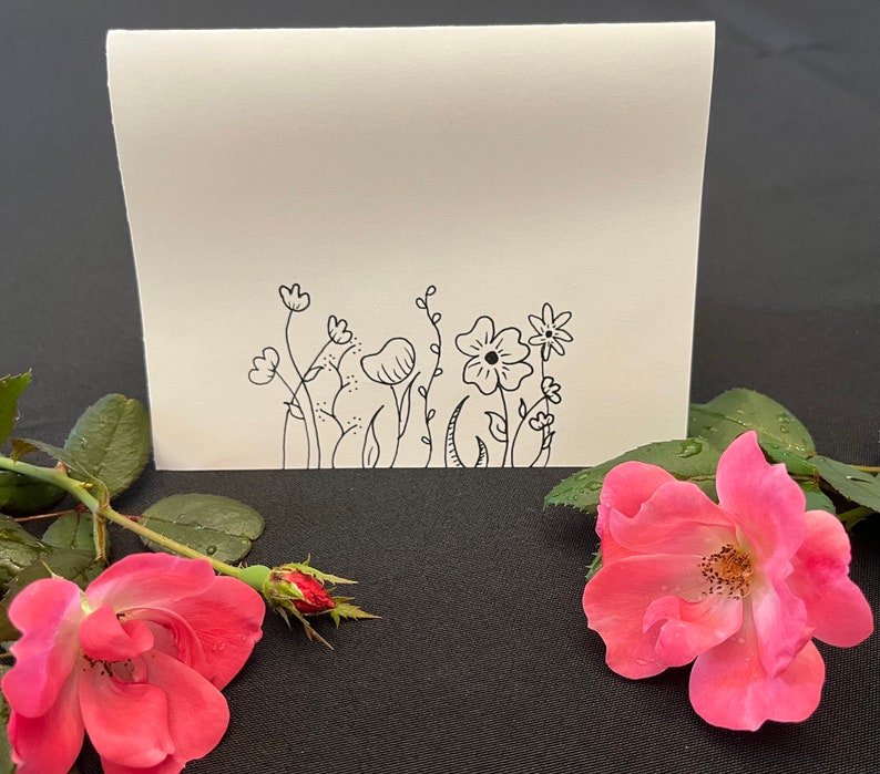 Puede incluir: Una tarjeta blanca en blanco con un dibujo de tinta negra de una fila de flores y tallos simples. La tarjeta est&aacute; sobre una superficie negra con una rosa rosa.