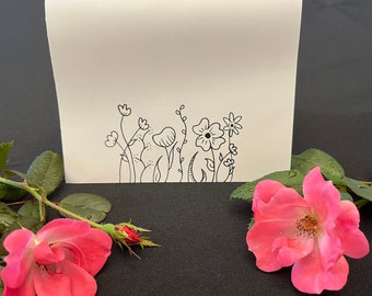 Puede incluir: Una tarjeta blanca con un dibujo lineal negro de flores y follaje estilizados en el borde inferior. Dos rosas rosas con gotas de agua a cada lado de la tarjeta, sobre un fondo negro.