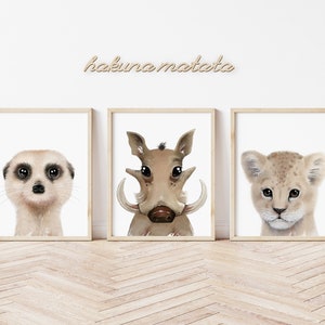 Può includere: Tre stampe di animali incorniciate con sfondo bianco. Le stampe mostrano un suricato, un facocero e un cucciolo di leone. Le cornici sono in legno marrone chiaro. Il testo "hakuna matata" è scritto in un carattere marrone chiaro sopra le stampe.