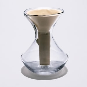 May include: Clear glass hourglass-shaped coffee maker with a light beige filter. The coffee maker has a wide top, a narrow middle, and a rounded base. The filter is filled with coffee grounds. The coffee maker sits on a white surface.