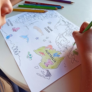 May include: A child coloring a world map with colored pencils. The map features illustrations of marine life and animals on the continents. The word "Australia" is visible on the map. Several colored pencils are on the table.