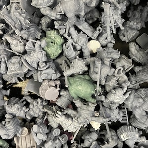 May include: A collection of unpainted miniature figurines, primarily in shades of gray, with a few green pieces. The figures appear to be fantasy characters, including warriors and creatures. The miniatures are scattered, suggesting a hobby or game.
