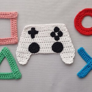 May include: A set of five crocheted items including a white game controller with black buttons, a pink square, a red circle, a green triangle, and a blue X-shaped cross. Each piece is crafted with visible yarn stitches and arranged on a flat surface.
