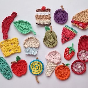 May include: A selection of crocheted food items in various colours, such as a red banana, green leaf, cake slice, purple plum, and a pie slice. Other items include a cupcake, pear, watermelon slice, strawberry, and orange. The items are arranged on a white surface.