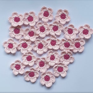 May include: A collection of 20 crocheted pink flowers with a red centre. The flowers are arranged in a loose cluster on a white background.
