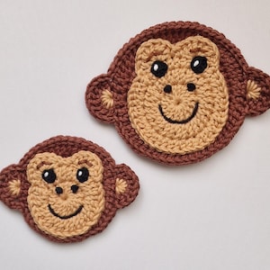 May include: Two circular crochet monkey face appliques in different sizes. Each piece features a tan face with dark brown outer borders, small ears, black embroidered eyes, a nose, and a curved mouth. The yarn texture is visible throughout.