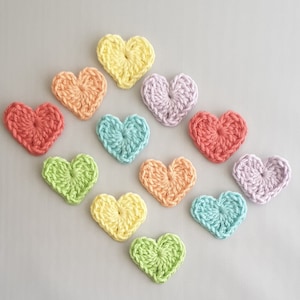 May include: A collection of 11 crocheted hearts in various pastel colours, including yellow, orange, red, pink, purple, blue, and green. The hearts are arranged in a diamond shape on a light grey background.