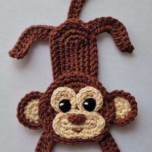 May include: A crocheted monkey toy in brown and beige. The monkey has a curved tail, arms, and legs. The face features large black eyes, a beige muzzle, and ears. This handmade item is suitable for nursery decor or as a playful gift for children.