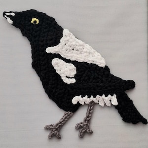 May include: A crocheted magpie appliqué featuring a black body with white wing patches and a white tail accent. The bird has a yellow eye with a black pupil and a white beak tip. Two grey crocheted legs with detailed feet extend from the bottom.