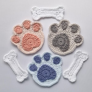 May include: A collection of crocheted paw prints and bone shapes. The paw prints are in pink, grey, and blue, with white outlines. The bones are white. These handmade items are likely pet-themed decorations or accessories.