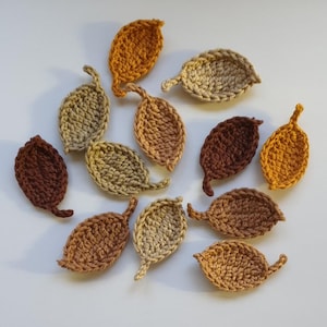 May include: A collection of twelve crocheted leaf appliques in various autumn-inspired colours including shades of brown, tan, beige, and mustard yellow. Each leaf features a textured stitch pattern and a small stem, arranged in a scattered layout.