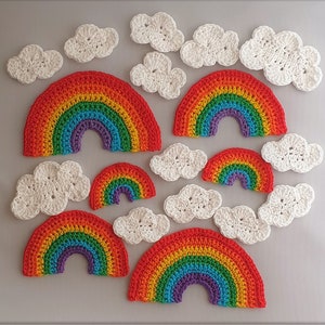 May include: A set of crocheted rainbow and cloud decorations. The rainbows are multi-coloured with red, orange, yellow, green, blue, indigo, and violet stripes. The clouds are white.