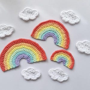 May include: Three crocheted rainbow appliqués in varying sizes, each featuring bands of red, orange, yellow, green, blue, and purple yarn. Surrounding the rainbows are five small, white, crocheted cloud appliqués with textured surfaces.