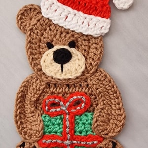 May include: A crocheted brown teddy bear appliqué wearing a red and white Santa hat. The bear has a cream-coloured muzzle with a black nose and holds a green gift box with a red ribbon and silver metallic thread accents on its lap.