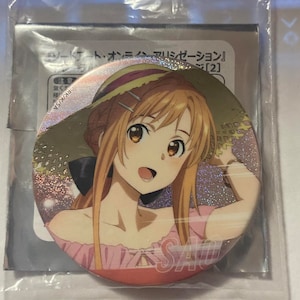 May include: A round, metallic button featuring a colorful anime illustration of a character with long, light brown hair and a straw hat. The character is wearing a pink top and the letters "SAO" are visible.