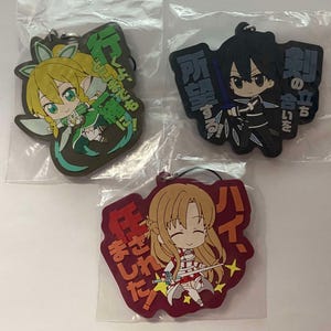May include: Three anime-style rubber keychains in plastic packaging. Each keychain features a different character in a chibi style. The keychains have Japanese text and are in various colors, including green, black, and red.