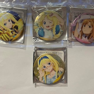 May include: Four round anime character badges in plastic packaging. Each badge features a different female character with blonde or brown hair. The badges have a glossy finish and are in various colors, including gold, pink, and blue.