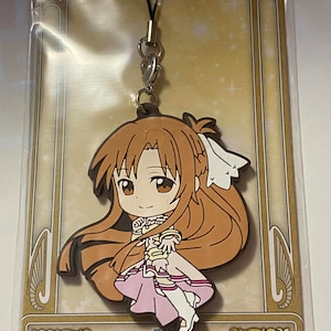 May include: A rubber strap featuring an anime character with long brown hair and a pink dress. The strap is attached to a black cord and a metal clasp. The packaging includes Japanese text and the Bandai logo.