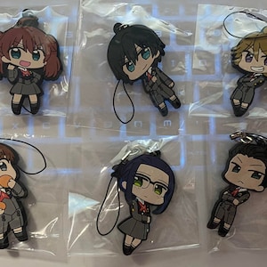 May include: Six anime-style character charms in clear plastic packaging. Each charm features a different character in a grey school uniform with red accents. The charms have black lanyards attached for hanging.