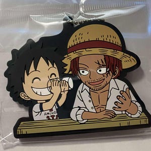 May include: A black rubber keychain featuring two cartoon characters. One character is wearing a straw hat and white shirt, the other is smiling and holding a cup. The keychain has a silver chain.