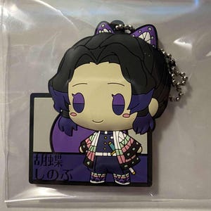 Puede incluir: Un llavero de goma con un personaje de dibujos animados con pelo negro y detalles morados. El personaje viste un atuendo estampado y tiene una espada pequeña. El llavero incluye una cadena de metal y texto japonés.