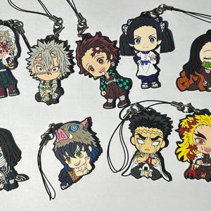 May include: A collection of ten colorful anime character charms with attached black lanyards. The charms feature various characters with distinct hairstyles and outfits, including a character with a boar head and another with a bamboo muzzle.