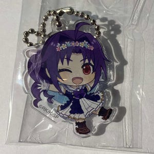 Puede incluir: Un colgante de acrílico transparente con un personaje de dibujos animados con cabello morado, una corona de flores y una expresión guiñando un ojo. El personaje viste un vestido azul oscuro con detalles blancos y botas marrones. Una cadena de bolas plateada está adjunta.